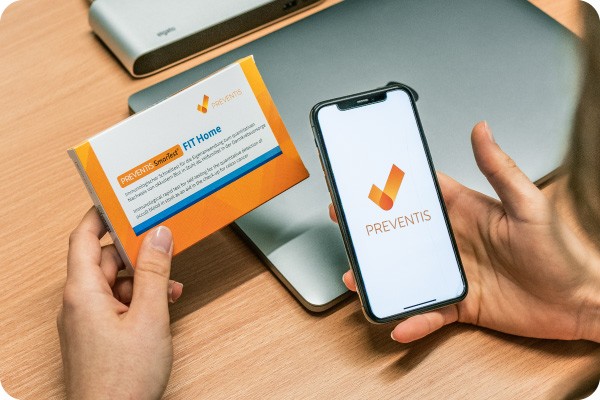 Person haelt den Schnelltest SmarTest FIT zur Darmkrebsvorsorge und ein Smartphone in der Hand.
