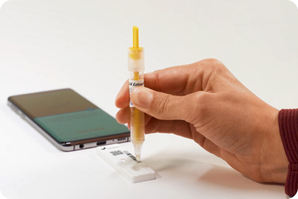 A close-up showing how a stool sample is applied to a test cassette for colorectal cancer screening, with a smartphone next to it.