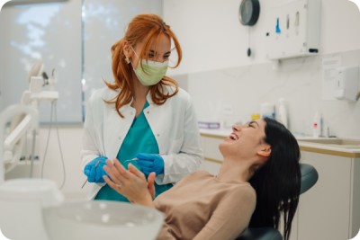 Weiblicher Zahnarzt mit chirurgischer Maske, die zahnaerztliche Werkzeuge haelt und mit gluecklicher Patientin spricht, die im Zahnarztstuhl in einer hellen modernen Zahnklinik sitzt.